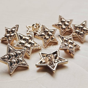 Peut inclure: Un ensemble de boutons en métal doré en forme d'étoiles à cinq branches. Chaque bouton présente une bordure perlée texturée et un motif floral à quatre pétales en relief au centre. Les boutons incluent une attache à boucle au dos.