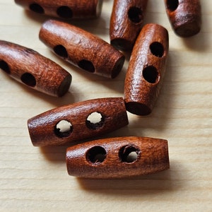 May include: A close-up of ten brown wooden toggle buttons, each with two holes. The buttons are arranged randomly on a light brown wooden surface.