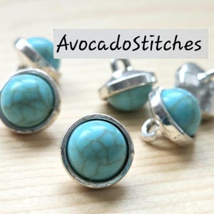 May include: Several round buttons featuring a smooth, turquoise-colored dome with brown veining, set within a polished silver-toned metal rim. Each button includes a small metal loop on the back for attachment. The text AvocadoStitches is visible.