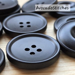 Peut inclure: Gros plan de plusieurs boutons noirs mats à quatre trous chacun. Les boutons sont ronds avec un bord légèrement surélevé. Le texte "AvocadoStitches" est dans le coin supérieur droit. Ces boutons sont idéaux pour les projets de couture et d'artisanat.