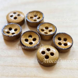 Peut inclure: Sept boutons ronds en bois, chacun avec trois ou quatre trous. Les boutons sont brun clair avec un bord brun plus foncé, disposés sur une surface en bois clair. Le texte "Avocadostitches" est visible sur certains boutons.