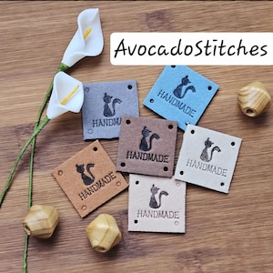 May include: Six square labels in blue, grey, brown, tan, cream, and white colours, each featuring a black cat silhouette and the text HANDMADE. The labels have four corner holes. Two white calla lily flowers and three wooden beads are present.