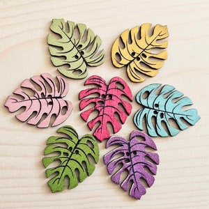 May include: Seven wooden monstera leaf buttons in various colours, including pink, green, blue, purple, and yellow. The buttons are all the same size and shape, and they have a hole in the centre for sewing.
