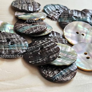 May include: A collection of round, two-hole buttons crafted from iridescent abalone shell. The buttons display a mix of dark, textured brown surfaces and smooth, pearlescent light surfaces with natural color variations and a glossy finish.