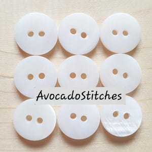 May include: A set of nine round, white, two-hole buttons arranged in a three-by-three grid. Each button has a smooth, slightly iridescent surface. The text "AvocadoStitches" is overlaid in black across the center of the button collection.