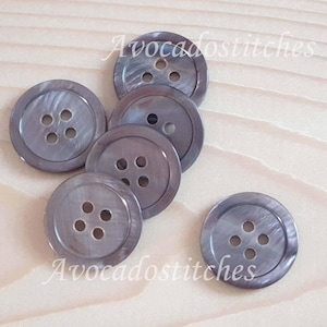 May include: Six grey mother of pearl buttons with four holes each. The buttons have a slightly iridescent sheen.