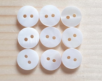May include: A set of nine circular white buttons arranged in a three-by-three grid. Each button features a smooth, iridescent surface with a subtle pearlescent sheen and two small, centered holes for threading, providing a clean and uniform appearance.