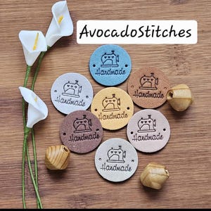 May include: A collection of seven circular labels in various colours including blue, tan, brown, and grey. Each label features a stamped graphic of a sewing machine with hearts and the word Handmade. The text AvocadoStitches is visible.
