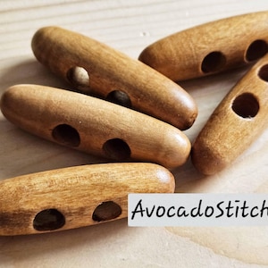 May include: Five light brown wooden toggle buttons, each featuring two circular holes for attachment. The buttons have a smooth, polished finish with visible wood grain patterns. A white rectangular label displays the text AvocadoStitches.