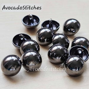 May include: A collection of eleven polished gunmetal grey shank buttons with a smooth, rounded dome shape. Each button features a reflective metallic finish and a small loop on the underside for attachment. The text AvocadoStitches is visible.