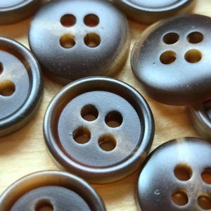 May include: A collection of round, dark brown buttons, each featuring four central holes. The buttons have a smooth, slightly glossy finish with subtle tonal variations in the brown colour, creating a depth that mimics natural horn or wood textures.