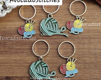 May include: Five metal stitch markers with circular rings and enamel charms. Three charms feature a ball of yarn with knitting needles in red, yellow, and blue. Two charms feature a larger ball of yarn in shades of green. Text reads AvocadoStitches.