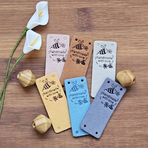 May include: A collection of six rectangular labels in various colours including light pink, tan, cream, yellow, blue, and grey. Each label features a stamped black graphic of a bee and flowers with the text "handmade with love" and two holes.
