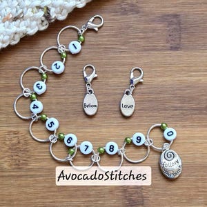 May include: A set of silver stitch markers with white numbered beads and green accents. Two charms read "Believe" and "Love". A charm with the text "follow your heart" is also included. The text "AvocadoStitches" is at the bottom.
