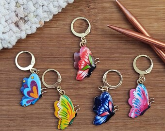 May include: Five colorful butterfly stitch markers with gold rings. The butterflies are painted in shades of blue, pink, red, yellow, and purple.