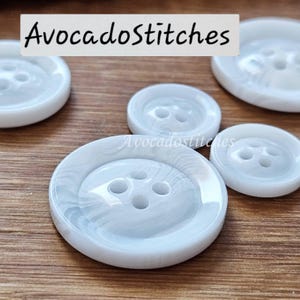 May include: A collection of round, four-hole buttons featuring a white and light grey marbled pattern. The buttons have a smooth, glossy finish and vary in size. The text "AvocadoStitches" is visible in the upper portion of the image.