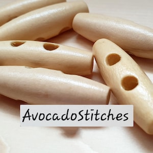 May include: A collection of light-colored, smooth, cylindrical wooden toggle buttons, each featuring two holes for attachment. A white rectangular label with the black text AvocadoStitches is positioned in the center of the image.
