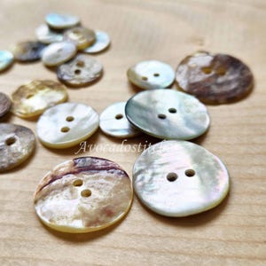May include: A selection of iridescent mother-of-pearl buttons in various sizes and shades of white, cream, and brown. Each button has two holes for sewing. The buttons are scattered on a light brown wooden surface.