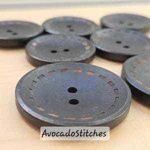 Peut inclure: Gros plan sur plusieurs boutons ronds gris foncé avec deux trous chacun. Chaque bouton a une bordure décorative cousue de couleur marron clair. Le texte "AvocadoStitches" est visible en bas.