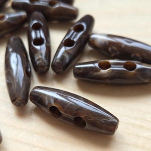May include: A collection of dark brown toggle buttons with a glossy, marbled finish. Each elongated button features two centred holes for attachment, showcasing a smooth, polished surface with subtle white swirling patterns throughout the material.