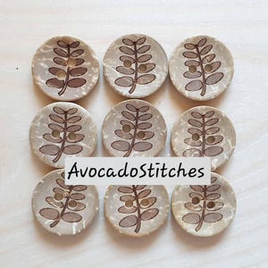 May include: Nine round, light beige buttons arranged in a three-by-three grid. Each button features a dark brown, laser-engraved leaf pattern with two central holes. The text AvocadoStitches is overlaid across the center of the image.