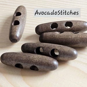 May include: Five dark brown wooden toggle buttons, each featuring two circular holes for attachment. The buttons have a smooth, elongated, cylindrical shape with rounded ends. The text AvocadoStitches is visible in the upper right corner.