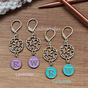 May include: Three gold-tone stitch markers with a leafy design and a round charm with a letter. The charms are in lavender and turquoise.