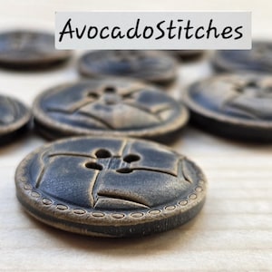 May include: A collection of round, dark gray buttons with a four-hole center and a decorative embossed border. Each button features a textured surface with a weathered finish. The text "AvocadoStitches" is visible at the top of the image.