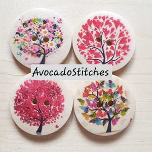 May include: Four round white buttons, each featuring a two-hole centre and a printed tree design. The designs include a floral tree, a heart-leaf tree, a pink-leaf tree, and a butterfly-leaf tree. The text AvocadoStitches is centred over them.