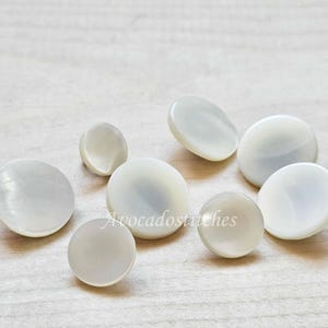 May include: A collection of eight round, smooth, white mother-of-pearl buttons of varying sizes. Each button features a glossy, iridescent finish with a slightly domed surface. The text Avocadostitches is overlaid in white across the centre.