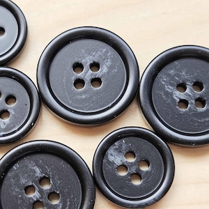 May include: Several black buttons with four holes, displayed on a light wooden surface. The buttons are of varying sizes and have a shiny finish. These are classic buttons for sewing projects.