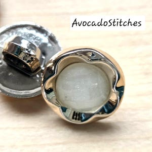 May include: Close-up of two decorative buttons. One button has a silver base and a gold-colored, scalloped edge. The center of the button is a light, opaque color. The other button has a silver base and a gold-colored, hexagonal loop. The text "AvocadoStitches" is in the image.