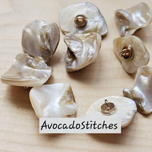 Peut inclure: Une collection de boutons en nacre irisée aux formes irrégulières et organiques avec une finition brillante. Chaque bouton est doté d'une attache en métal doré au dos. Le texte AvocadoStitches est visible sur l'image.