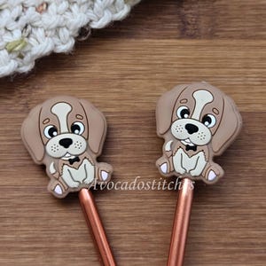 May include: Two copper knitting needles with brown and white dog-shaped toppers. The dogs have black eyes, brown noses, and white bow ties.
