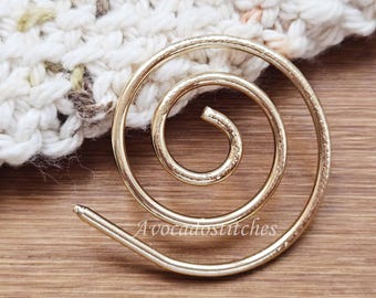 May include: A gold-toned metal spiral pin with a smooth, polished finish. The circular wire design coils inward toward the center. The text "AvocadoStitches" is printed in white across the lower portion of the gold spiral metal accessory.