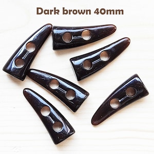 May include: Six dark brown horn-shaped buttons with two holes each. The buttons are 40mm long and are arranged in a scattered pattern on a light wood surface.