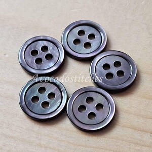 May include: Five circular dark grey buttons with a subtle iridescent sheen and a smooth finish. Each button features four holes arranged in a square pattern in the centre. The text Avocadostitches is overlaid across the centre of the buttons.