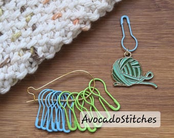 May include: A collection of knitting accessories, including blue and green stitch markers, a gold safety pin, and a yarn-themed charm. The accessories are arranged on a wooden surface, with a portion of a knitted item visible in the background. The text "AvocadoStitches" is also visible.