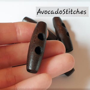 May include: A set of dark brown wooden toggle buttons with two holes for sewing. Each button has a cylindrical, elongated shape with rounded ends and a smooth, polished finish. The text AvocadoStitches is visible in the upper right corner.