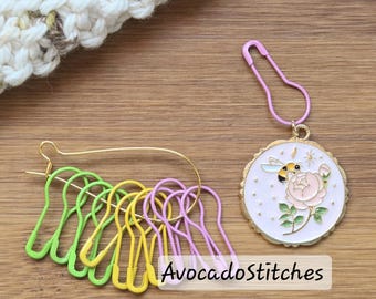 May include: A collection of knitting stitch markers in green, yellow, and purple, along with a decorative charm featuring a bee and a rose. The charm is attached to a pink safety pin. The text "AvocadoStitches" is visible.