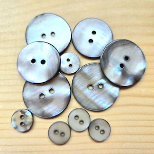 May include: A collection of ten round, iridescent grey buttons of varying sizes, each featuring two central holes. The buttons exhibit a smooth, polished surface with a subtle pearlescent sheen, displaying shades of charcoal, silver, and light grey.