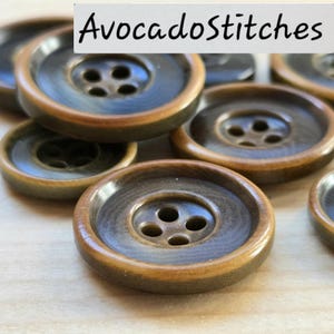 May include: A collection of round, four-hole buttons featuring a dark grey centre and a light brown outer rim. The buttons have a smooth, polished finish and are arranged in a group. The text "AvocadoStitches" is visible at the top of the image.
