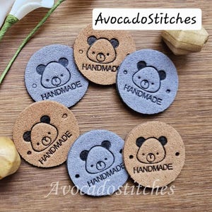 May include: A collection of circular tan and light blue leather tags featuring a stamped bear face graphic and the word HANDMADE. The tags have small holes on the sides for attachment. A white label with the text AvocadoStitches is visible.