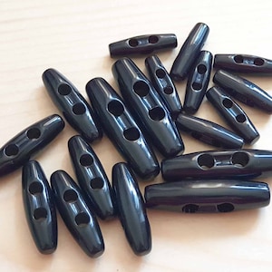 May include: A collection of black toggle buttons, each featuring a smooth, elongated cylindrical shape with two centered holes for attachment. These fasteners are uniform in color and have a glossy finish, arranged in a scattered group.