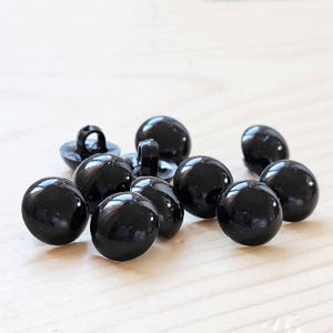 May include: A group of ten black plastic buttons. The buttons are round and have a shiny finish.