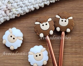 Può includere: Due segnapunti a forma di pecora marrone su spilli in metallo color rame e due segnapunti a forma di pecora bianca. Ogni pecora ha un muso color crema, occhi neri e un motivo di lana testurizzato. Il testo Avocadostitches è visibile.