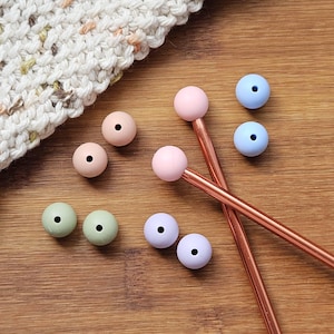 May include: Two copper-coloured knitting needles with round, pastel-coloured silicone toppers in pink and light blue. Several loose, spherical silicone beads in shades of pink, light blue, sage green, and lavender are arranged nearby.
