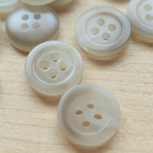 May include: A collection of round, off-white buttons, each featuring four central holes and a slightly recessed centre. These circular fasteners have a smooth, semi-translucent finish and are arranged in a cluster, showing their uniform design.