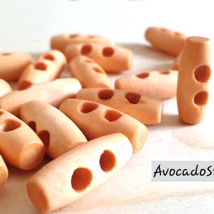 May include: A collection of light orange wooden toggle buttons, each with two holes. The buttons are approximately 20mm in length. The image includes the text "AvocadoStitches".