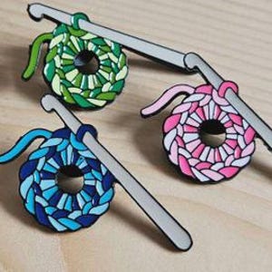 May include: Three enamel pins depicting crocheted circles with a crochet hook. The circles are green and white, pink and white, and blue and white.